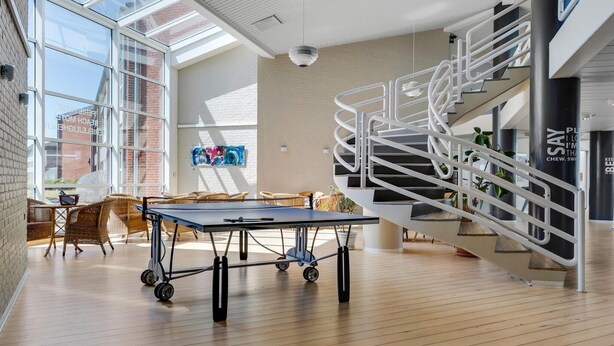 A spacious indoor area with a ping pong table, wicker furniture, a spiral staircase, and large windows allowing natural light. - Fanø Bad, værelse 23 (Fanø)