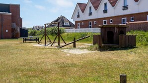 A playground with swings and a small wooden structure, surrounded by a grassy field and residential buildings with red-tiled roofs. - Fanø Bad, værelse 23 (Fanø)