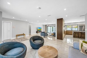 A modern living room with a sofa, armchair, and coffee table, a kitchen area with a dining table, and a large window.
