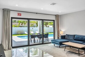 A modern hotel room with a large sliding glass door leading to a pool area, a blue sofa, and a wooden coffee table.