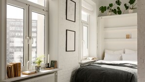 A bedroom with a large window, a radiator, a bed with a dark bedspread, and a shelf with books and a potted plant. - Lwowska Apartment (Warsaw)