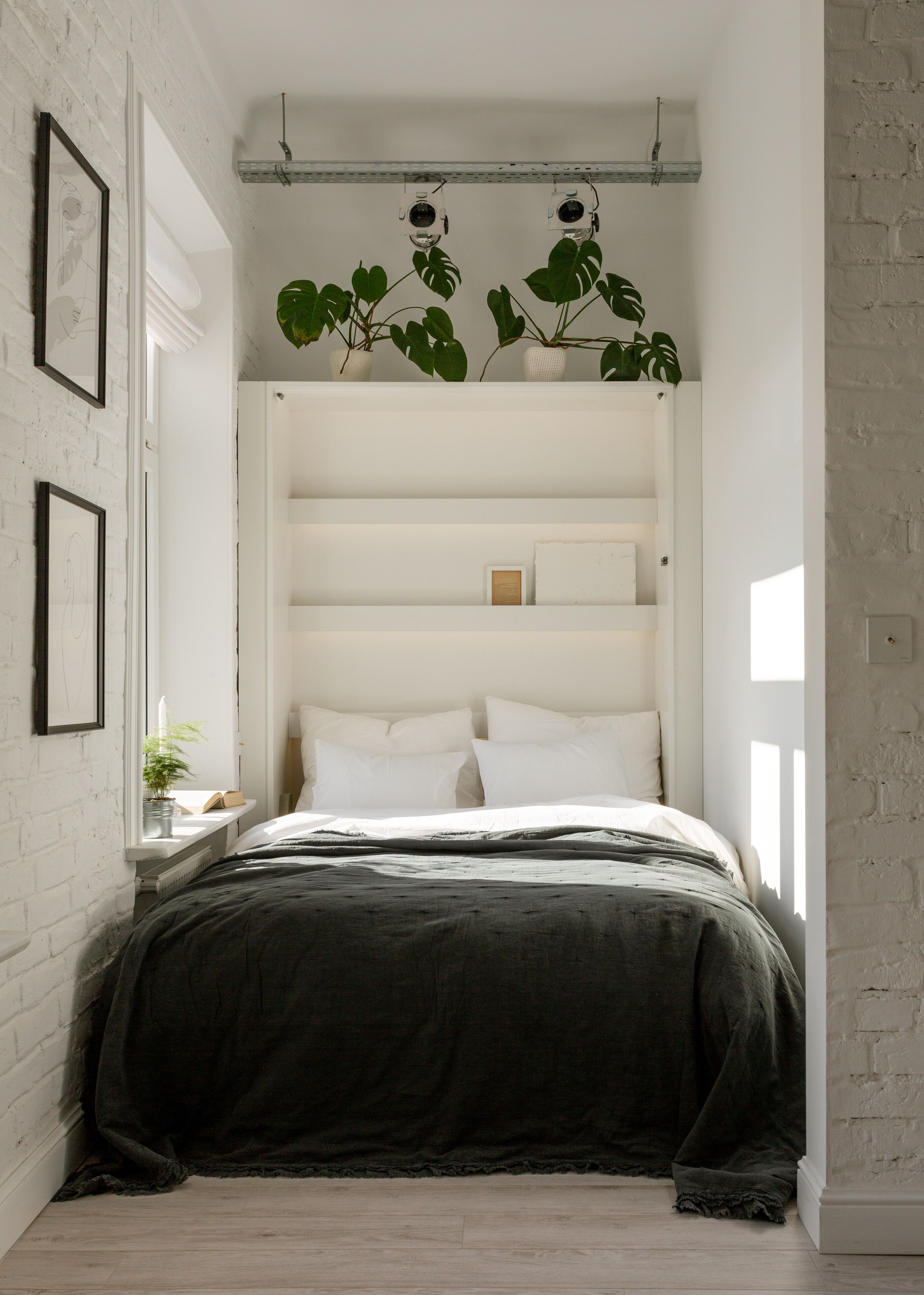 A bedroom with a bed, white walls, a shelf with plants, and a camera on the ceiling.