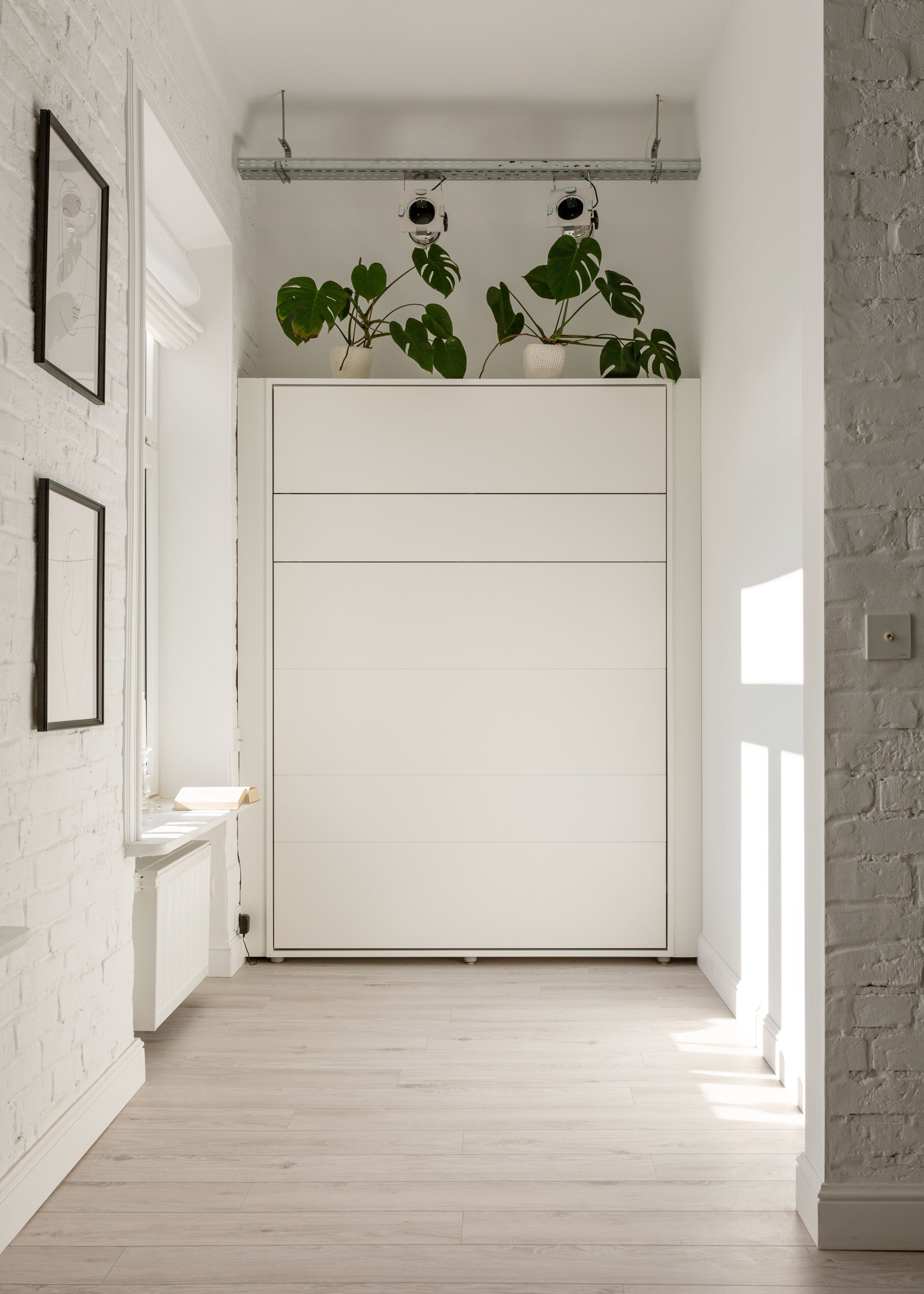 A hallway with a white door, white brick walls, and a radiator. There are two cameras mounted on the ceiling and a plant with large leaves on the wall.