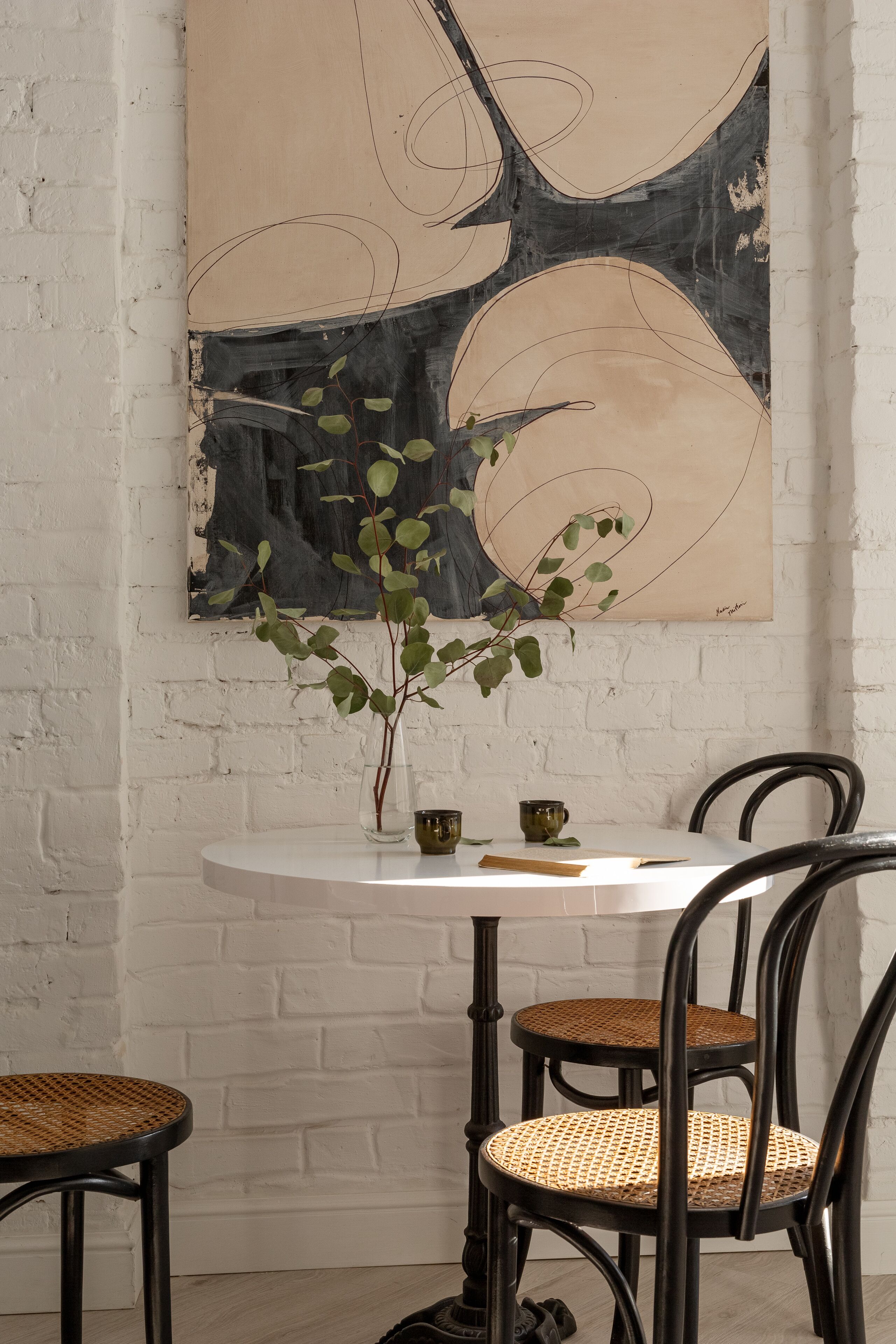 A round table with a vase of green leaves, two wicker chairs, and a wall-mounted abstract painting.