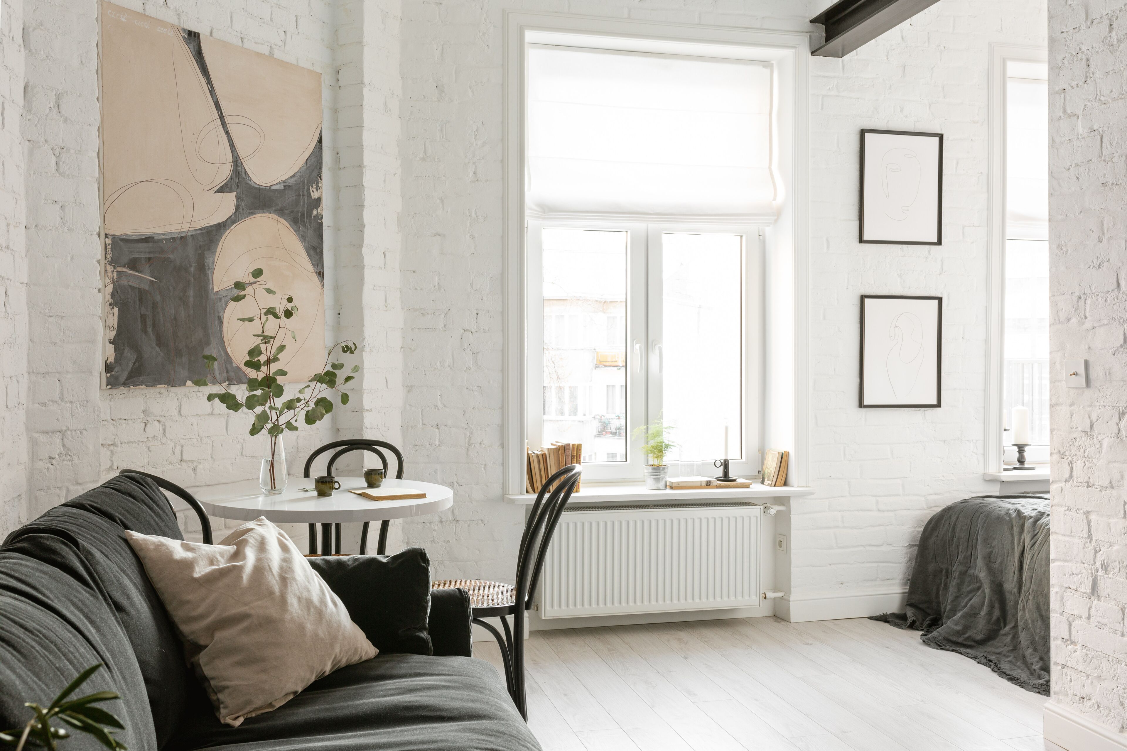 A modern living room with a white brick wall, a large window, a black sofa, a dining table, and a radiator.