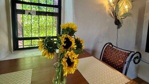 Une pièce avec une table en bois, un vase rempli de tournesols, une fenêtre donnant sur un paysage verdoyant et un luminaire fixé au mur.