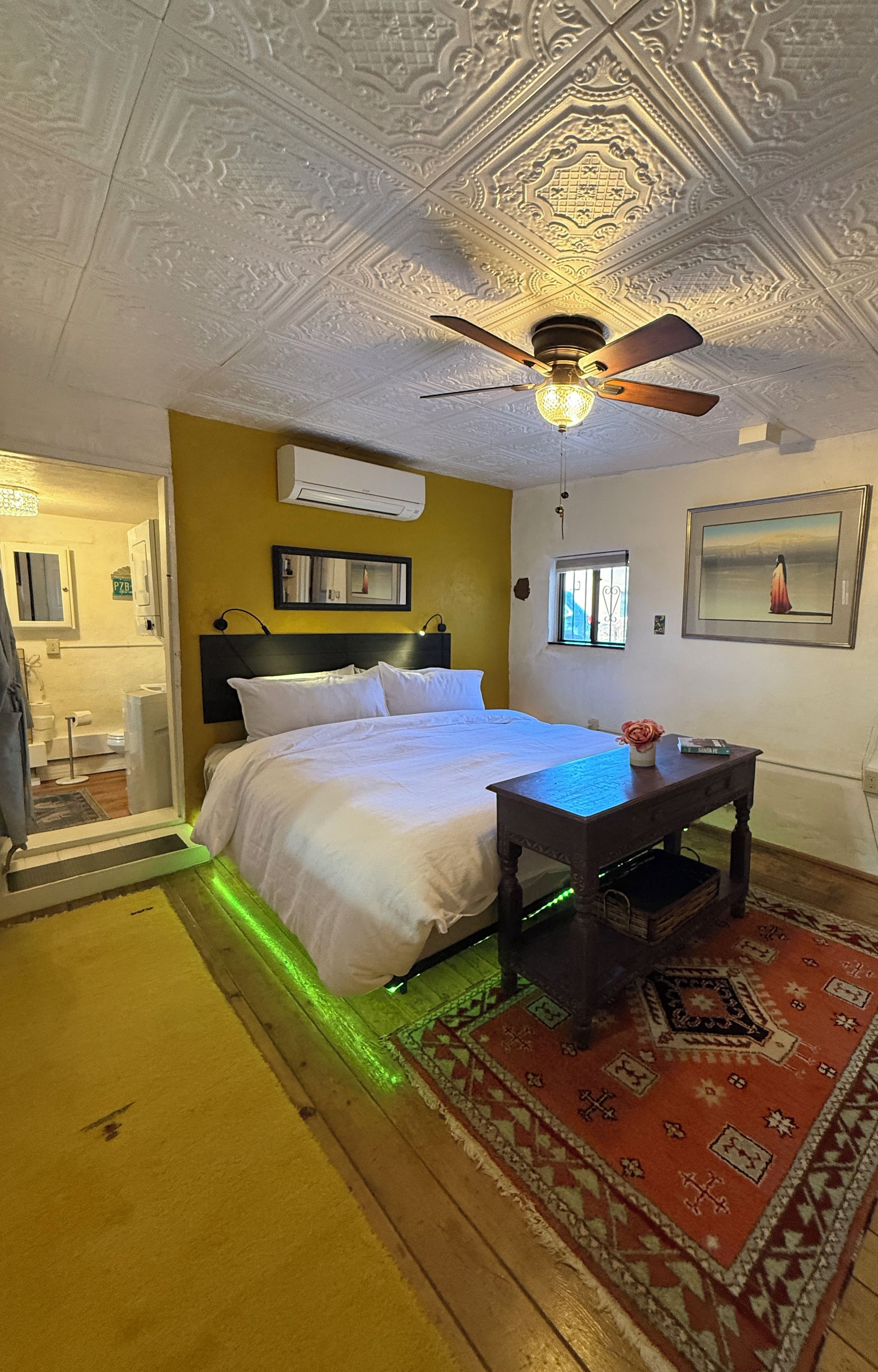 A hotel room with a bed, a ceiling fan, a table, a rug, and a painting on the wall.