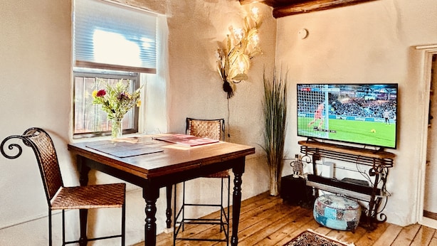 Un coin repas avec une table en bois, des chaises, un vase de fleurs, un téléviseur posé sur un meuble et une fenêtre avec des stores.