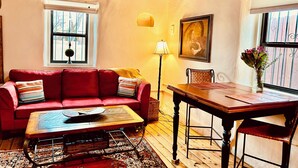 Un salon chaleureux avec un canapé rouge, une table basse en bois et une table à manger avec des chaises. On y trouve des fenêtres avec des rideaux, une lampe et un tableau au mur.