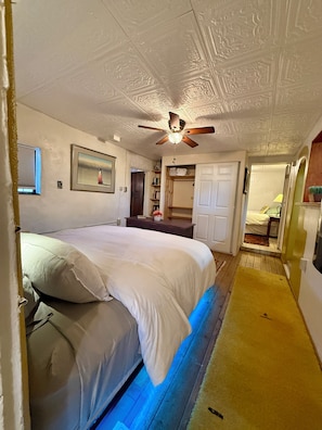 A bedroom with a bed, a ceiling fan, a framed picture, and a door leading to another room.
