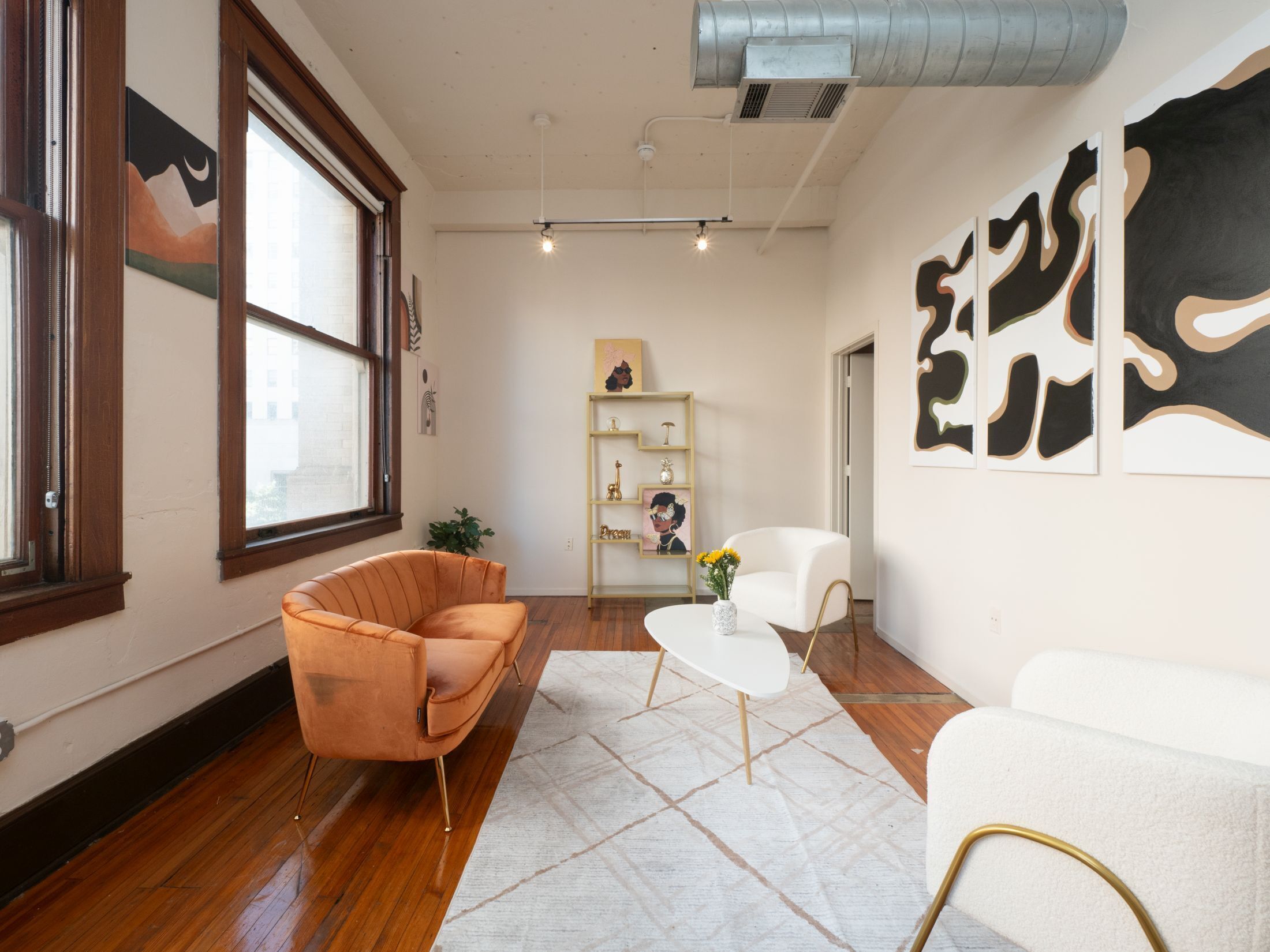 A modern living room with a wooden floor, a brown leather sofa, a white armchair, and a white coffee table. There is a bookshelf with decorative items and a large abstract painting on the wall.