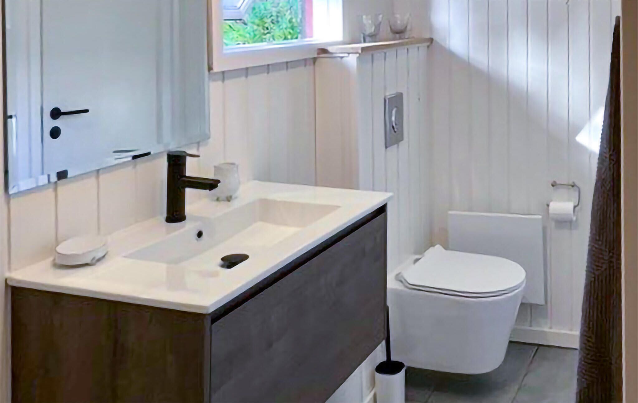 Een badkamer met een wit wastafelmeubel, een donkere onderkast, een wit toilet en een raam boven de wastafel.