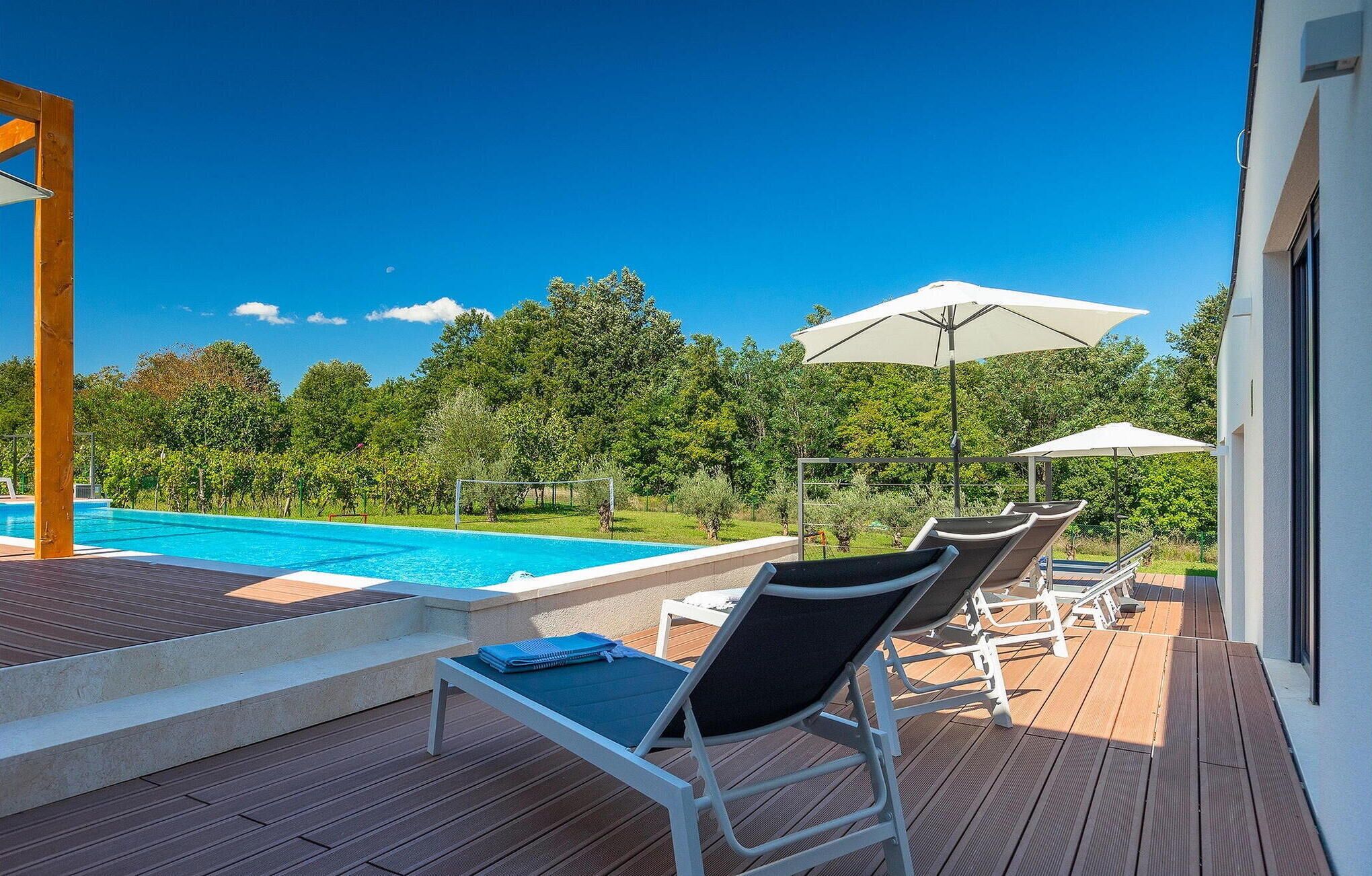 Ein Poolbereich mit Liegestühlen und Sonnenschirmen, ein Holzdeck und Blick auf einen Swimmingpool und Grünflächen.