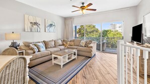 A spacious living room with a large sofa, a coffee table, and a TV on a wooden stand. There is a balcony with a railing and a view of greenery and buildings.