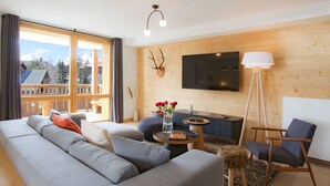 Una sala moderna con un sofá grande color gris, un tabique de madera, un televisor montado y vistas a un paisaje montañoso a través de amplias ventanas.