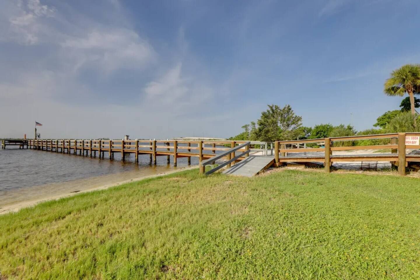 Une jetée en bois s’étendant sur des eaux calmes, avec une zone herbeuse au premier plan et un ciel dégagé.