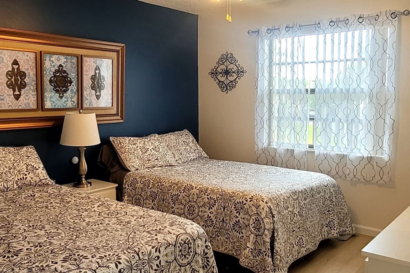 A room with two beds, a nightstand with a lamp, a decorative wall piece, and a window with patterned curtains.