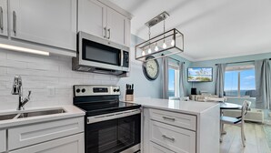 A modern kitchen with white cabinetry, a built-in oven, a microwave, and a dining area with a view of the outdoors. - Perdido Skye 71 | Beachfront, Pool, Private Balcony | My Beach Getaways (Pensacola)