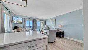 A modern kitchen with a white countertop, a dining area with a table and chairs, a grey sofa, and a small side table with a lamp. - Perdido Skye 71 | Beachfront, Pool, Private Balcony | My Beach Getaways (Pensacola)