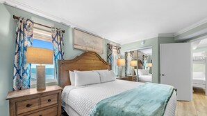 A bedroom with a wooden headboard, a large bed with a white quilt, a green throw blanket, a nightstand with a lamp, and a window with patterned curtains. - Perdido Skye 71 | Beachfront, Pool, Private Balcony | My Beach Getaways (Pensacola)