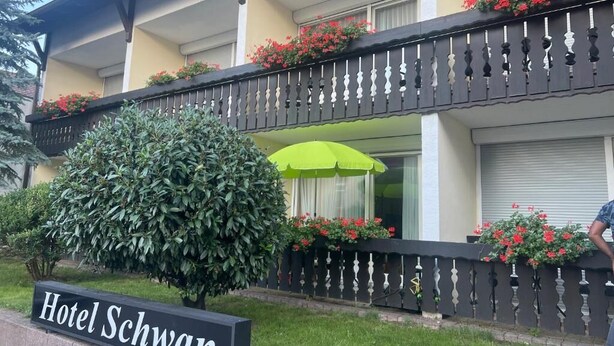 Et hotel med et skilt, der siger "Hotel Schwan". Der er en balkon med sort gelænder og røde blomster. En grøn parasol er placeret på balkonen.