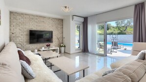 Un soggiorno moderno con una televisione a schermo piatto montata su una parete in mattoni, una porta in vetro che si apre su un'area piscina e un divano con cuscini soffici.