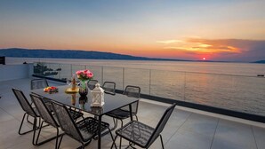 Une table à manger avec des chaises, sur une terrasse donnant sur un plan d’eau, au coucher du soleil.