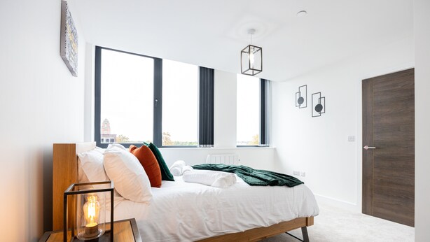 A modern bedroom with a wooden bed, large windows, a bedside table with a lamp, and a view of a building outside. - Chic Industrial Luxury Stay (Stretford)