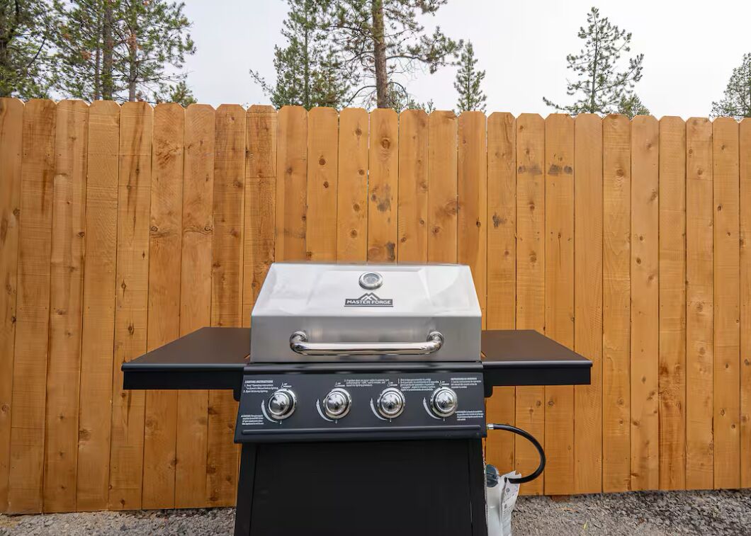 A MasterForce gas grill with four control knobs, set against a wooden fence backdrop.
