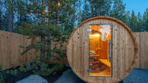 Una cabaña de madera con diseño circular, cuyo interior muestra una cama y una ventana, rodeada por una cerca de madera y árboles.
