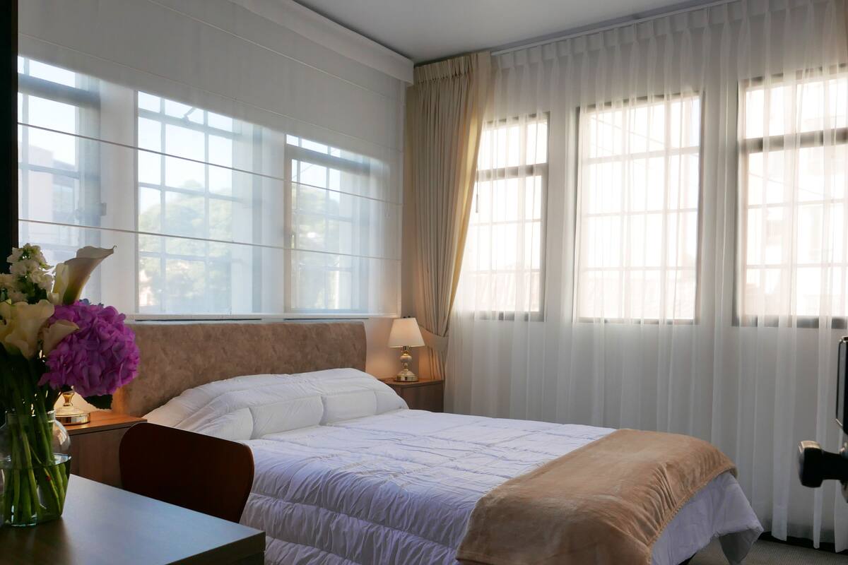 Ein Hotelzimmer mit Bett, Nachttisch mit Lampe, Blumen in einer Vase und einem Fernseher auf einem Schrank.