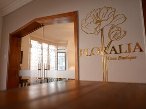 A room with a wooden floor, a glass door leading to another room, and a wall with a floral design and the text "FLORALIA Casa Boutique". - Floralia Casa Boutique (Bogotá)