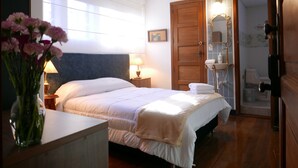 Ein Hotelzimmer mit einem großen Bett, einem Nachttisch mit Lampe, einer Holztür und einem durchgegangene Tür erkennbaren Badezimmer.