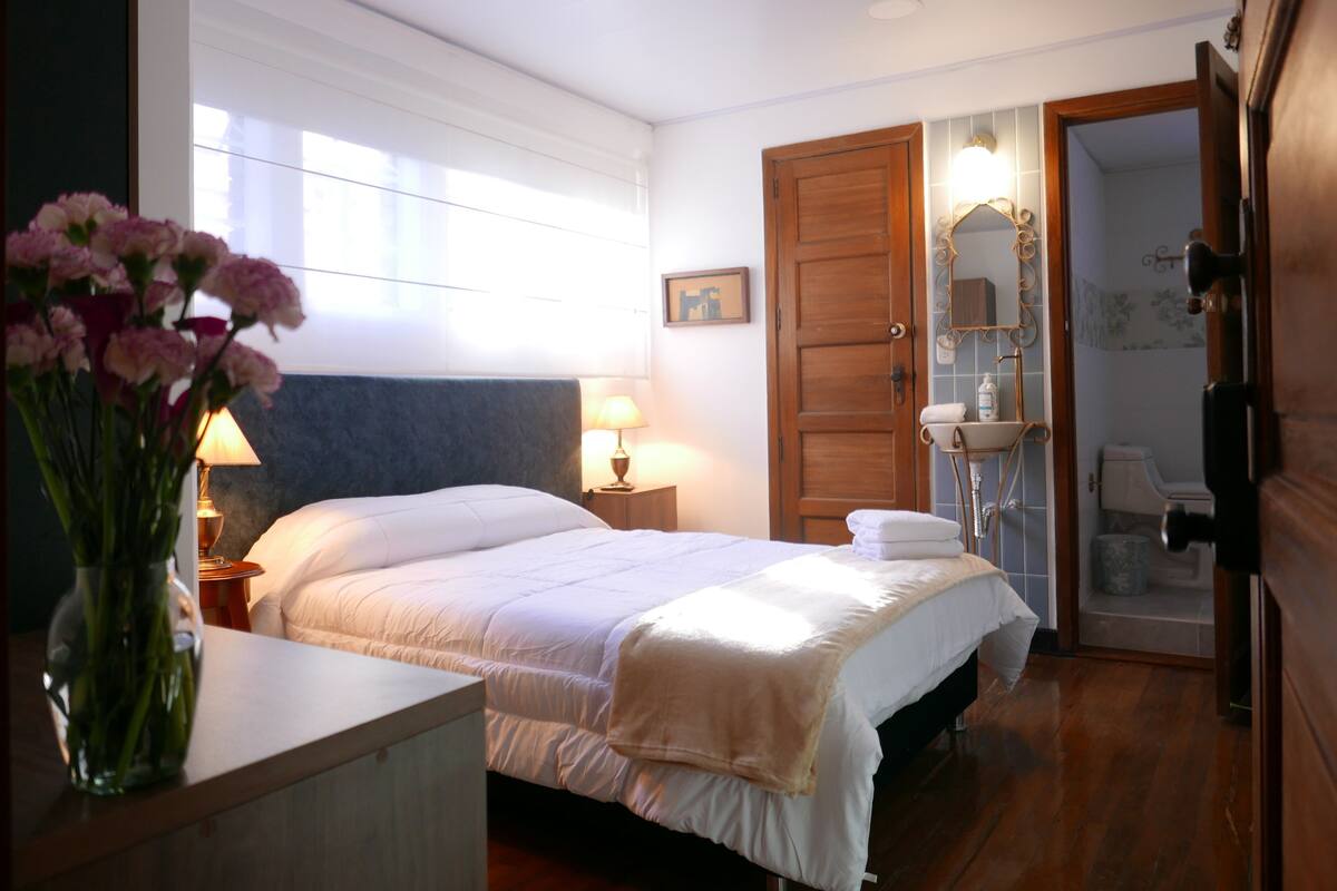 Ein Hotelzimmer mit einem großen Bett, einem Nachttisch mit Lampe, einer Holztür und einem durchgegangene Tür erkennbaren Badezimmer.