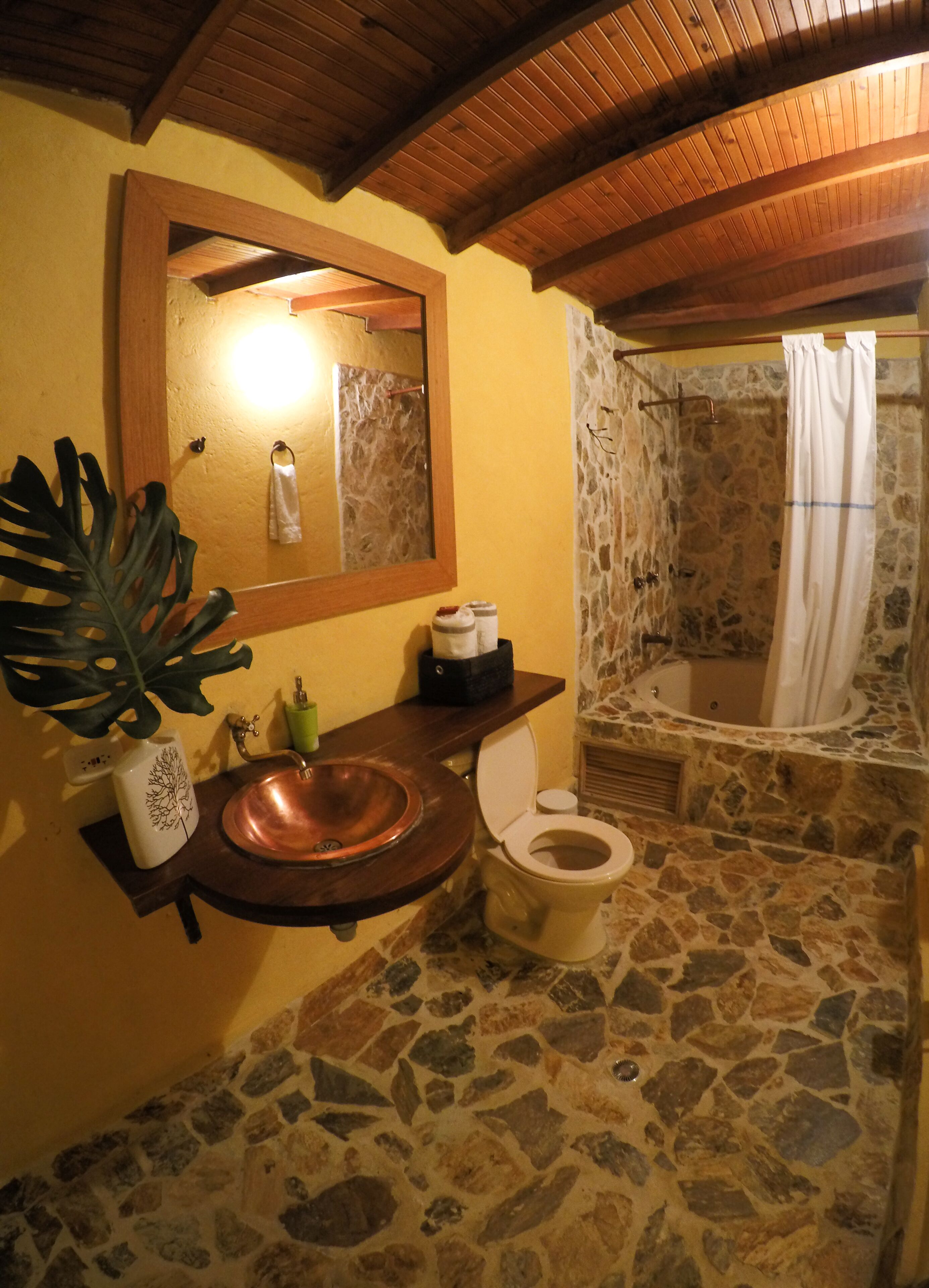 Un bagno con pavimento in pietra, un mobiletto in legno con lavandino in rame, un water bianco, uno specchio grande e uno spazio doccia con parete in pietra.