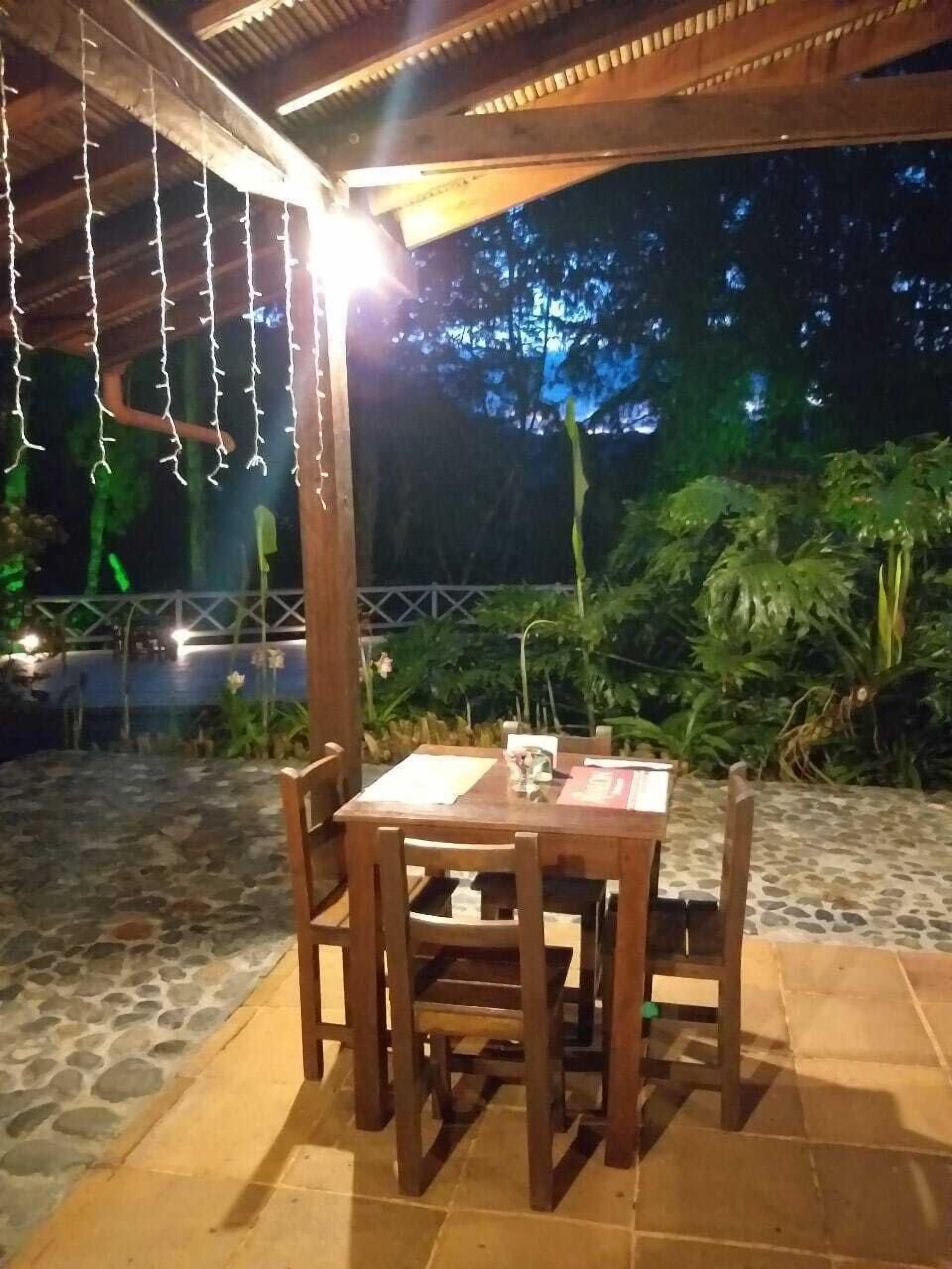 Mesa de madera al aire libre con sillas, rodeada de baldosas de piedra y follaje, bajo una pérgola de madera con luces colgantes.