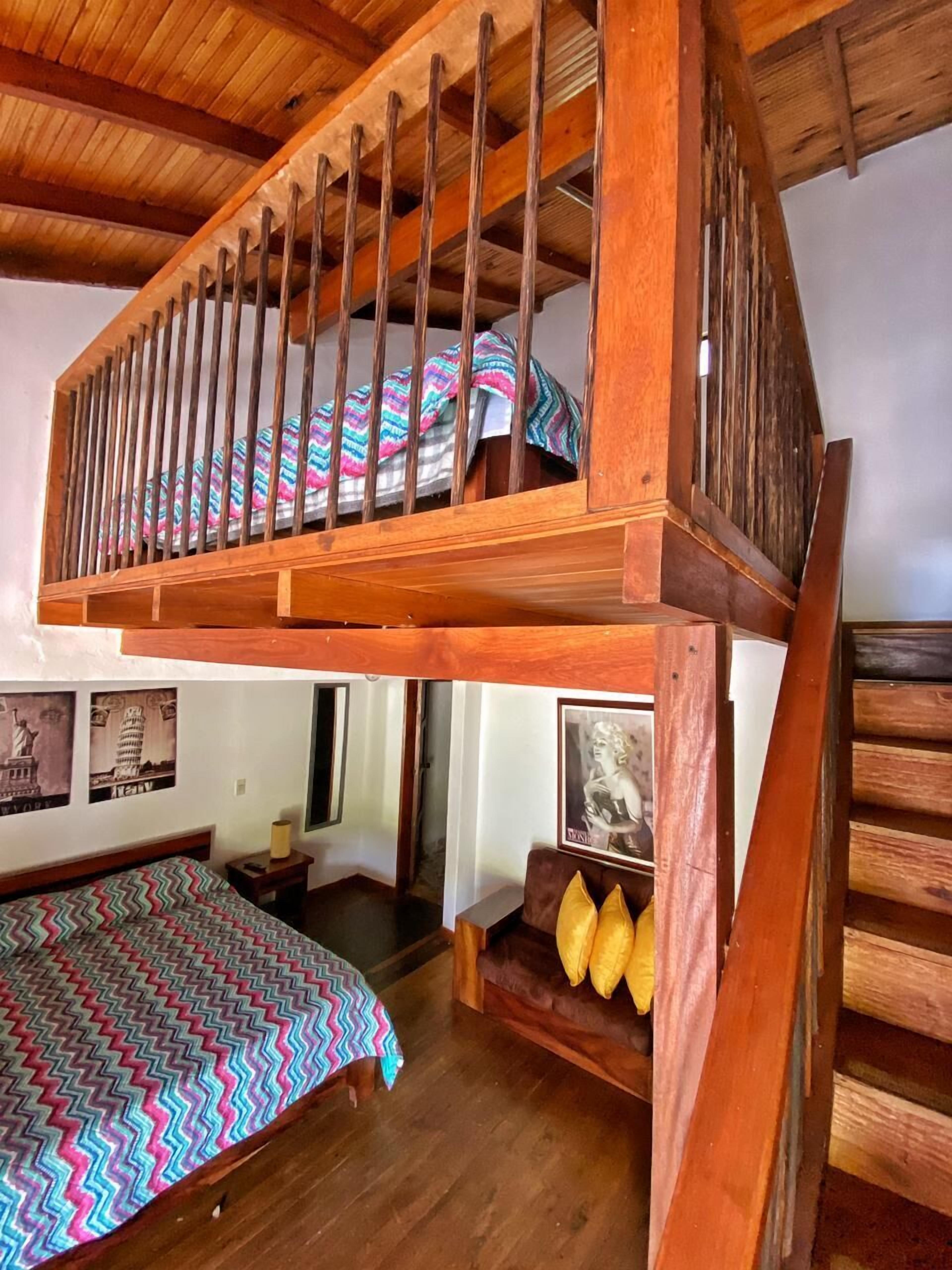 A room with a wooden bunk bed, a bed with a colorful zigzag bedspread, and a small seating area with cushions.