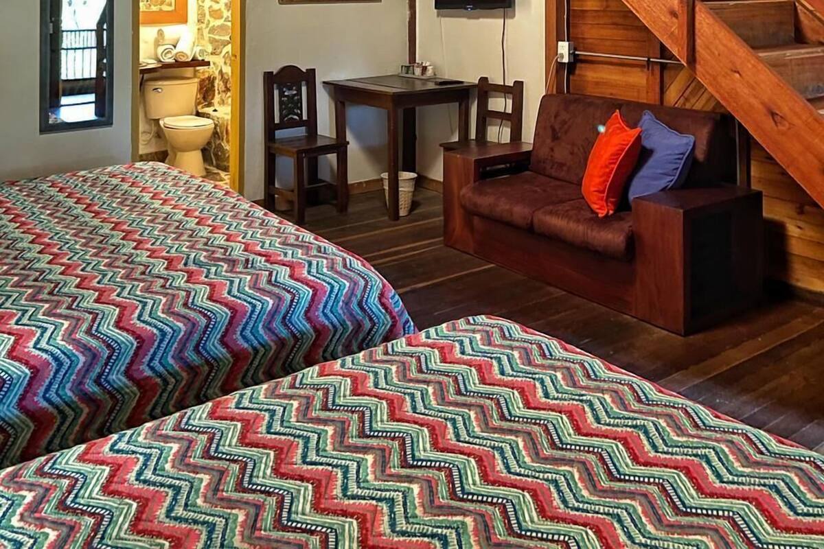 A rustic room with a wooden staircase, a bed with patterned bedding, a sofa with cushions, and a desk with a chair.