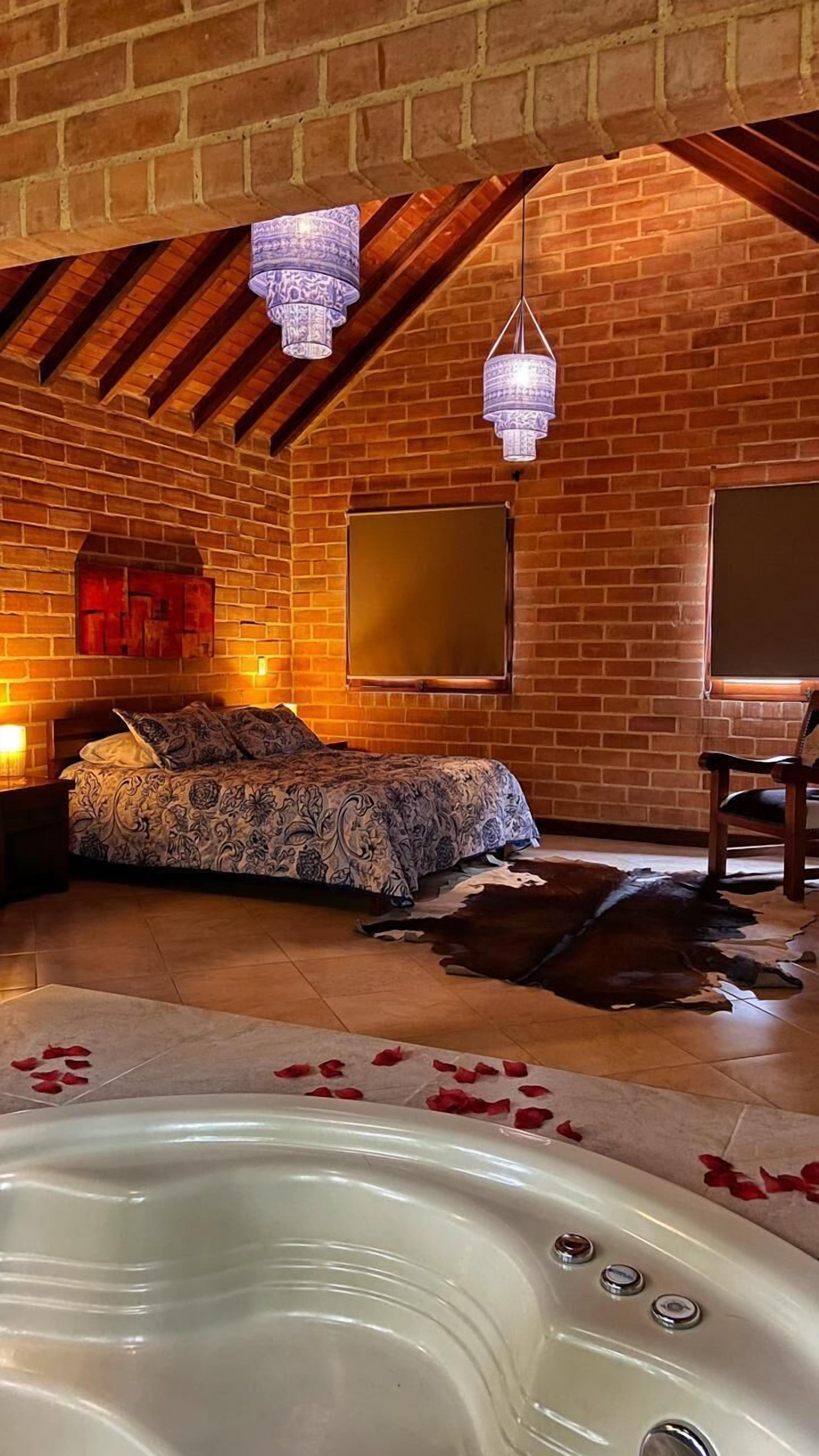 A room with a bed, a wooden chair, a large window, a brick wall, and a hot tub with rose petals.