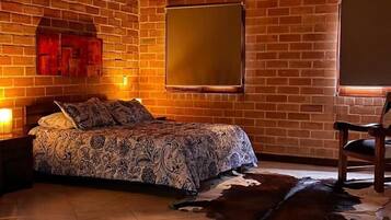 A room with a bed, a wooden chair, a large window, a brick wall, and a hot tub with rose petals.