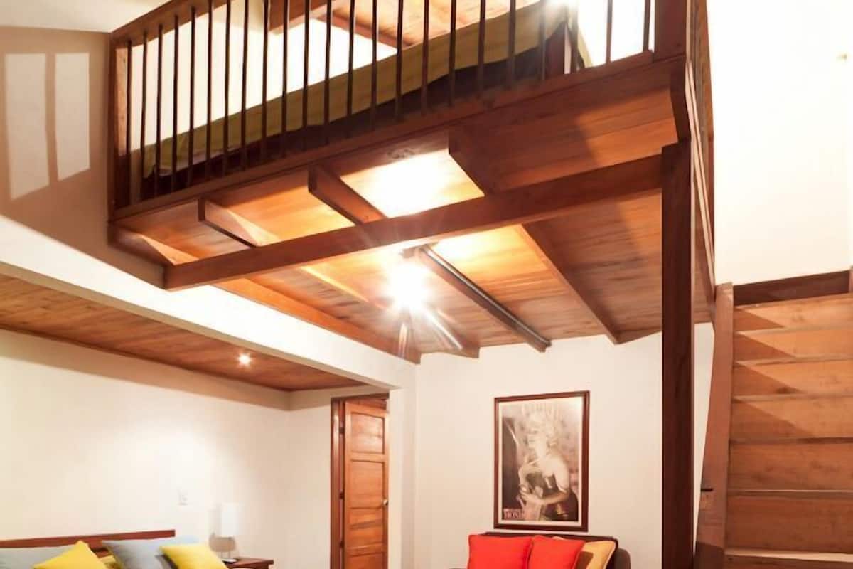 A room with a wooden staircase, a bed with yellow bedding, a brown sofa with red cushions, and a framed picture on the wall.
