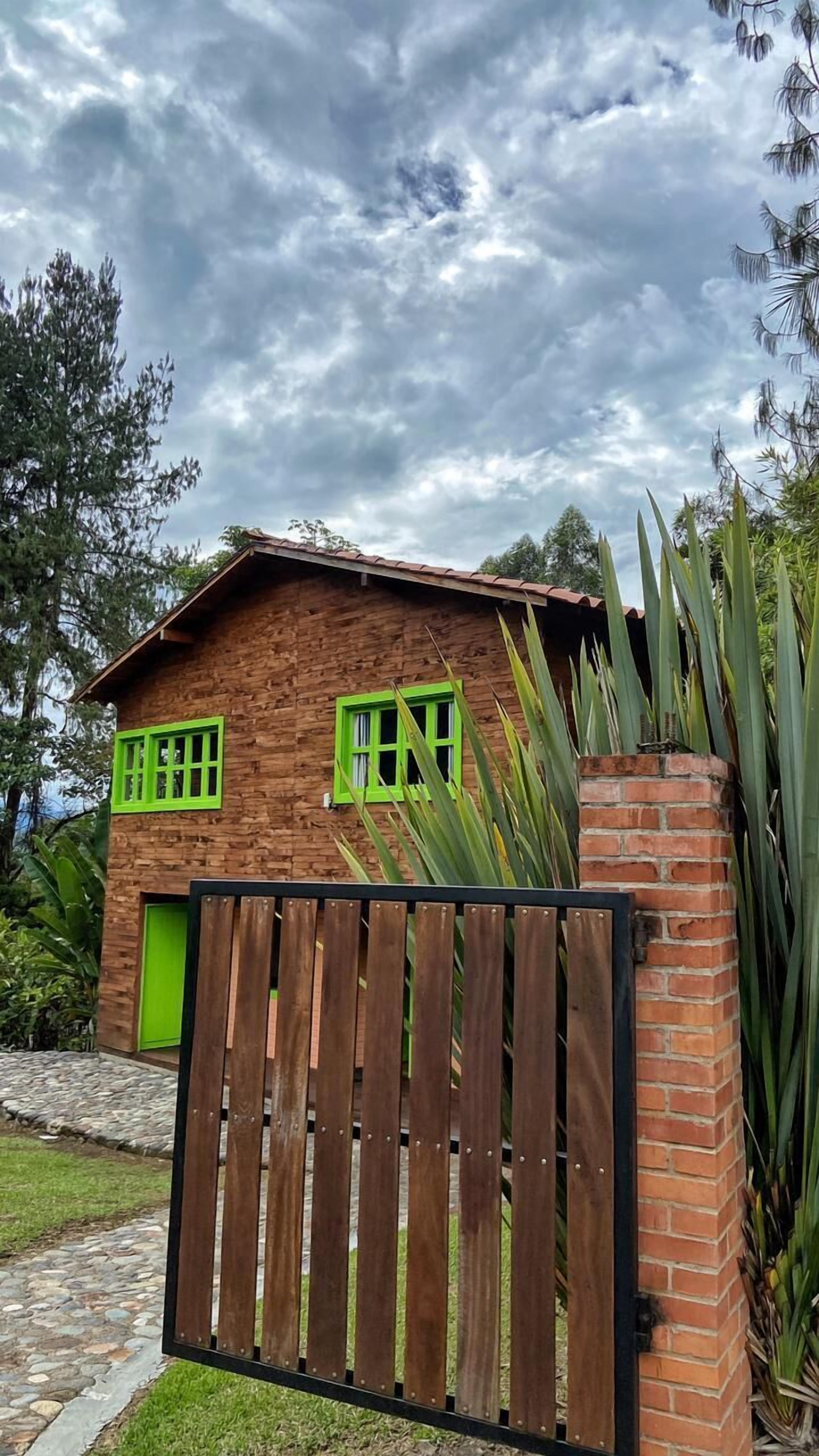 Una casa de madera con contraventanas verdes y un muro de ladrillo, rodeada de follaje.
