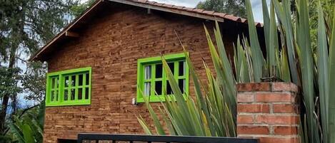 Una casa de madera con contraventanas verdes y un muro de ladrillo, rodeada de follaje.