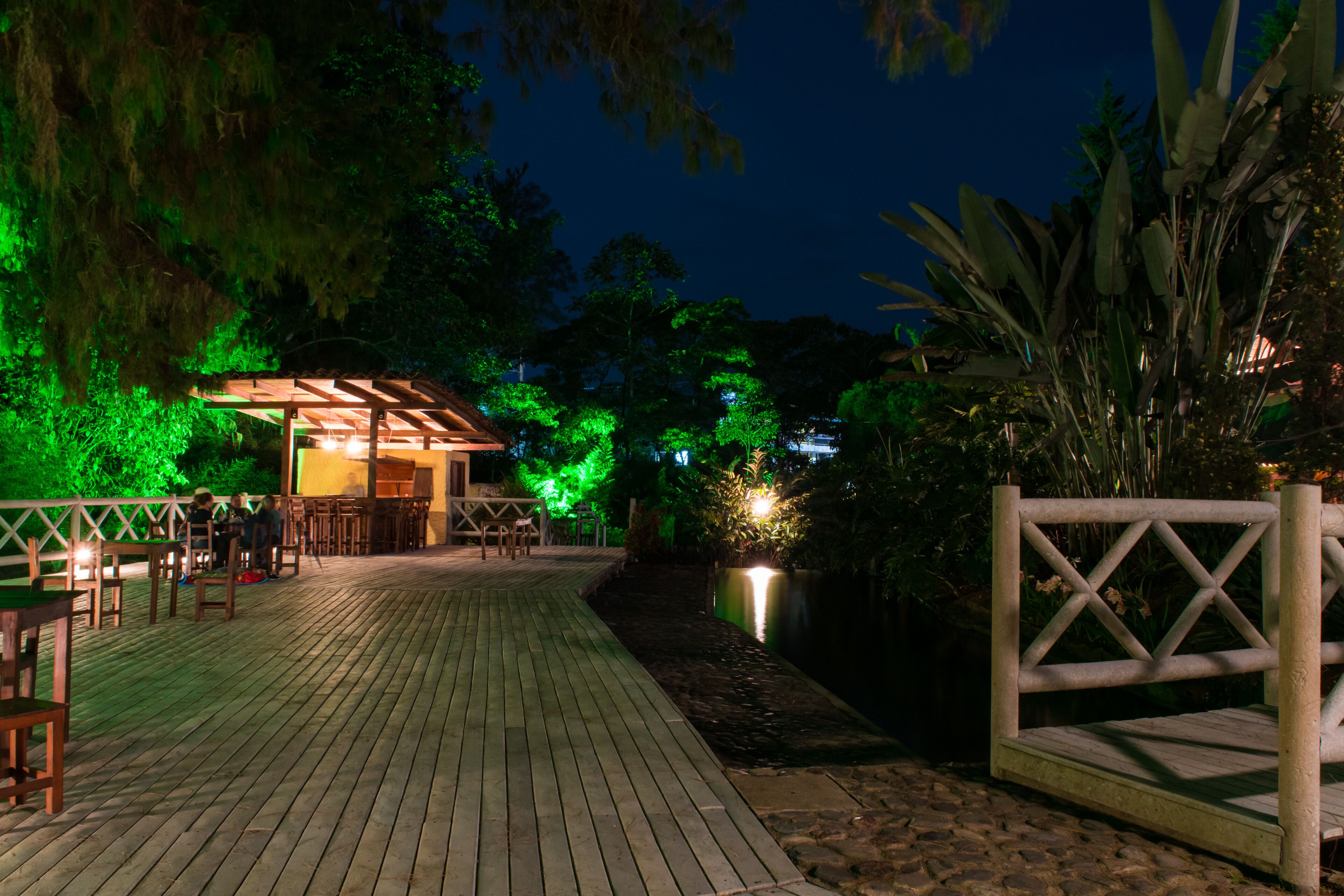 A wooden deck with chairs and tables, surrounded by greenery and a building with green lighting.