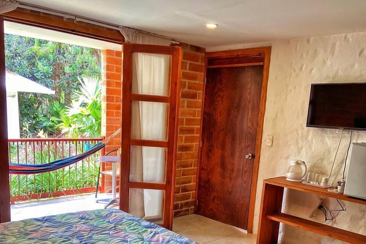 A bedroom with a bed, a TV, a small table with a kettle, and a balcony with a hammock.