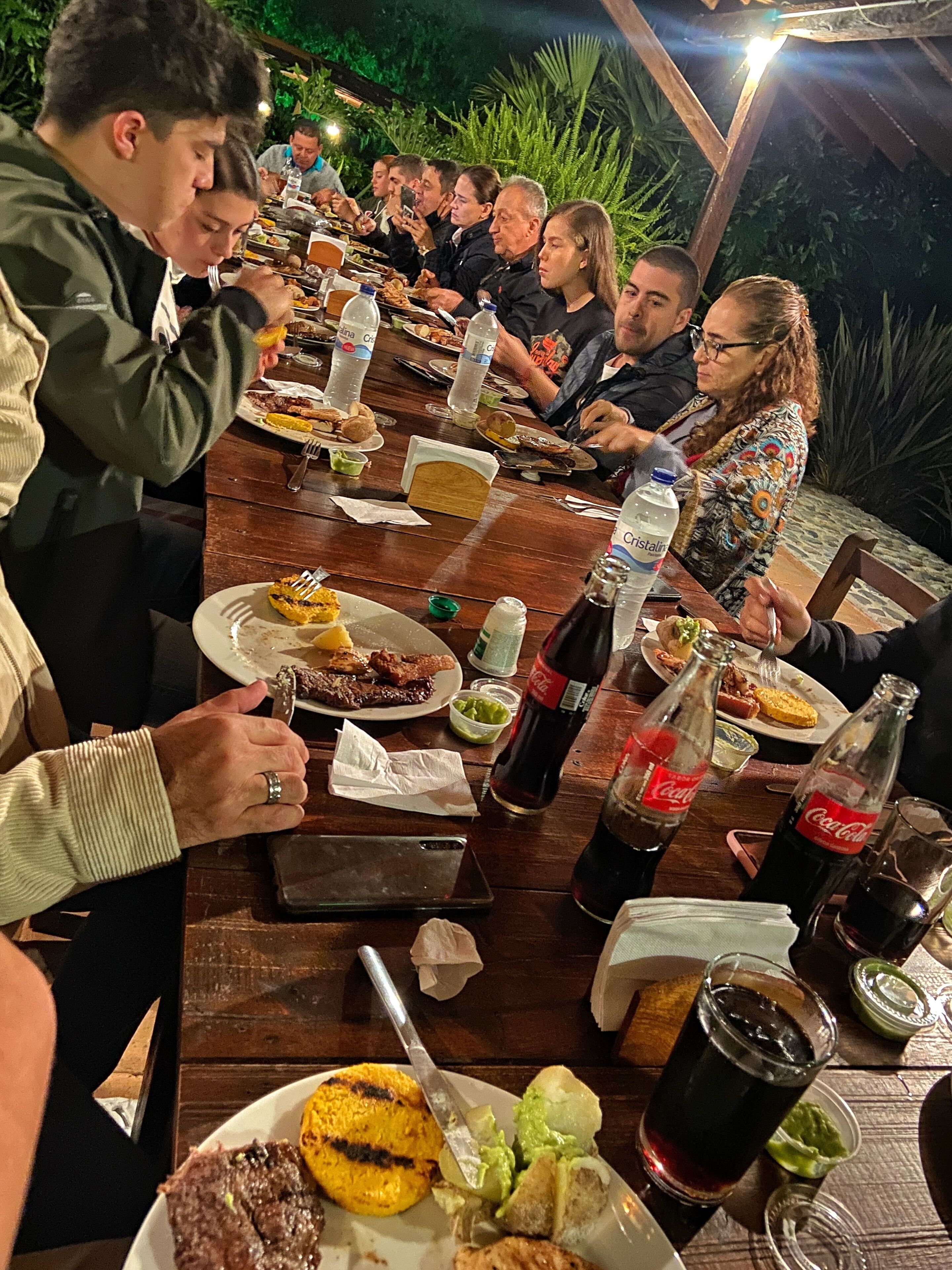 A group of people dining at an outdoor wooden table with various dishes and drinks.