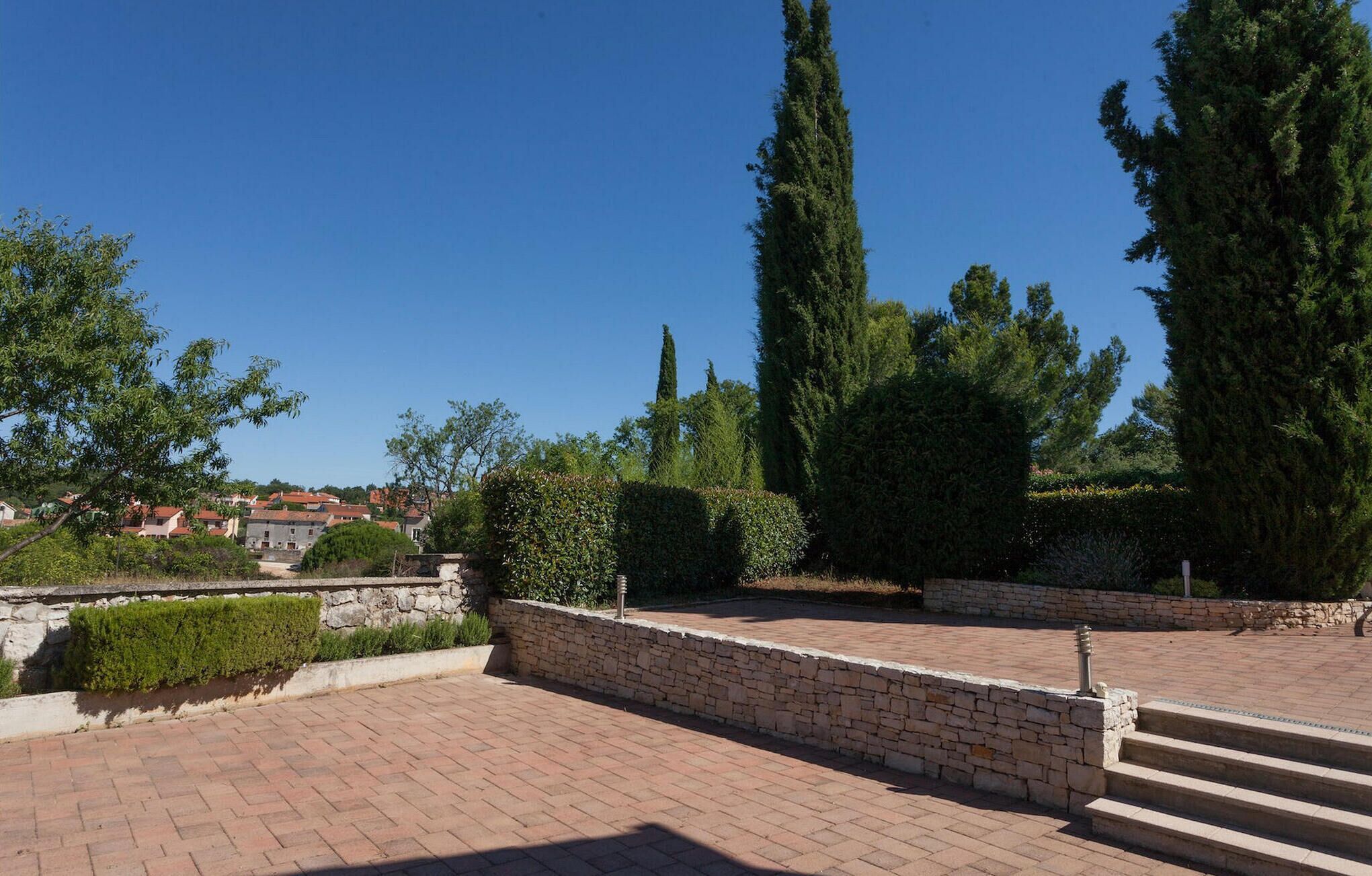 A paved courtyard with stone walls and steps leading up to a garden area with tall trees and shrubs.