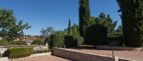 A paved courtyard with stone walls and steps leading up to a garden area with tall trees and shrubs.