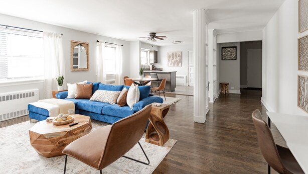 Un vaste salon avec un canapé bleu, une table basse en bois et des fauteuils en cuir marron. On aperçoit à l’arrière-plan un coin repas avec une table et des chaises blanches.