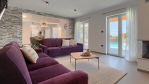 Ein modernes Wohnzimmer mit einer Wand aus Stein, einer violetten Sofagarnitur, einem Holztisch und Blick auf einen Swimmingpool durch große Glasfenstertüren.